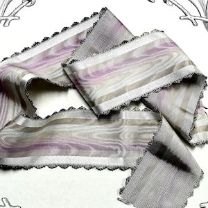 May include: A length of decorative ribbon with a striped pattern in shades of gray, lavender, and cream. The edges are finished with a delicate, scalloped trim. The ribbon is folded and draped, showcasing its texture and design.