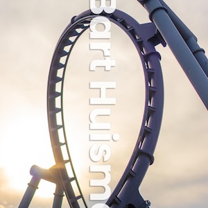 May include: A close-up of a roller coaster track against a cloudy sky. The track is made of metal and is painted a dark gray color. The track is in the shape of a loop, and the sun is shining through the loop.
