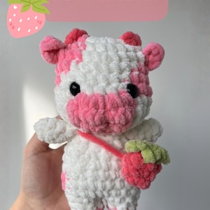Peut inclure: Une vache en peluche faite à la main, rose et blanche, tenant une fraise. La vache a des yeux noirs et une sangle rose. L'image comprend le texte "PDF PATTERN" et les drapeaux des États-Unis et de la France.