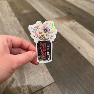 May include: A holographic sticker featuring a Coca-Cola Zero can with a bouquet of flowers. The can is black with the Coca-Cola Zero logo in white and red. The flowers include daisies and other wildflowers.