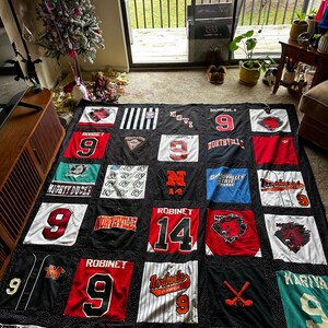 May include: A black and white quilt made from old t-shirts. The quilt features a variety of sports team logos and names, including "Mighty Ducks", "Northville", "Novi", "Robinet", "Kariya", and "Cool Story".