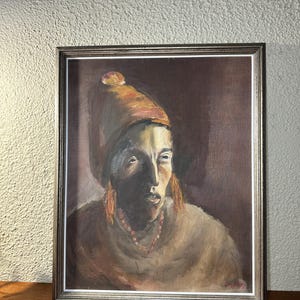 Puede incluir: Retrato al óleo de una persona con sombrero marrón y collar de cuentas. La pintura está enmarcada en un marco de color plateado y está sobre un fondo neutro. El rostro del sujeto está iluminado.