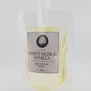 May include: A clear plastic pouch containing 100ml of light yellow reed diffuser refill liquid. The label features a circular logo with wavy lines, the text "WHITE MUSK & VANILLA", "REED DIFFUSER REFILL", and "100ml" printed in black on a light grey background.