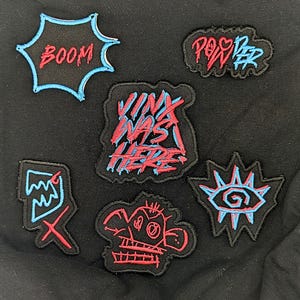 以下が含まれることがあります： 黒い背景に、赤と青のステッチで刺繍された6つのパッチのコレクション。パッチには様々なデザインとテキストがあり、「BOOM」、「POWER」、「Vinx Was Here」など。 他のパッチは様式化されたグラフィックを描いています。