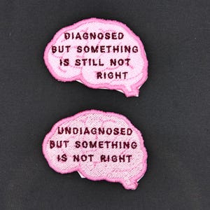 Può includere: Due toppe di feltro rosa a forma di cervello con testo ricamato. La toppa superiore recita "Diagnosticato ma qualcosa non va ancora". La toppa inferiore recita "Non diagnosticato ma qualcosa non va".