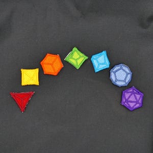 Op de afbeelding: Een regenboog van kleurrijke geborduurde pleisters in de vorm van dobbelstenen, waaronder een rode driehoek, een geel vierkant, een oranje kubus, een groene ruit, een blauwe ruit, een donkerblauwe dodecaëder en een paarse icosaëder.