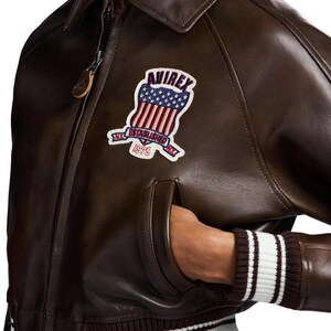 May include: Brown leather bomber jacket with a zip closure, a collar, and a patch on the chest. The patch features a shield design with stars and stripes, and the text "AVIREX NY ESTABLISHED 1975". The jacket has a ribbed cuff with white stripes.