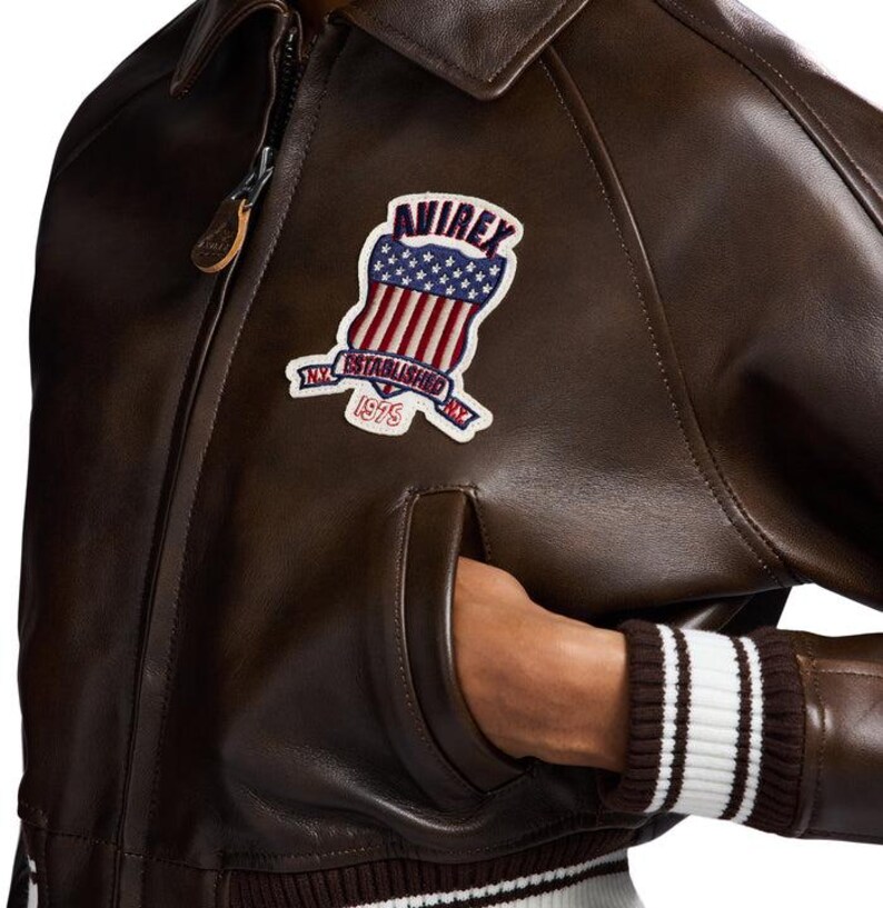 May include: Brown leather bomber jacket with a zip closure, a collar, and a patch on the chest. The patch features a shield design with stars and stripes, and the text "AVIREX NY ESTABLISHED 1975". The jacket has a ribbed cuff with white stripes.