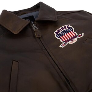 May include: Close-up of a brown leather jacket with a collar and a zipper. The jacket features a patch with the text "AVIREX ESTABLISHED 1975" and a design of the American flag.