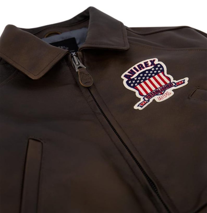 May include: Close-up of a brown leather jacket with a collar and a zipper. The jacket features a patch with the text "AVIREX ESTABLISHED 1975" and a design of the American flag.