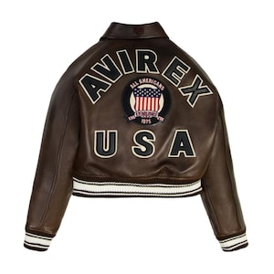 May include: A brown leather jacket with the word "AVIREX" across the shoulders and "USA" below. The jacket has a patch with the American flag and the words "ALL AMERICANS ESTABLISHED 1975". The jacket has white and brown striped cuffs and waistband.