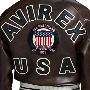 May include: Brown leather jacket with the word "AVIREX" across the shoulders in black and white lettering. A circular patch with the words "ALL AMERICANS" and "ESTABLISHED 1975 N.Y." is centered below the lettering. The word "USA" is below the patch.