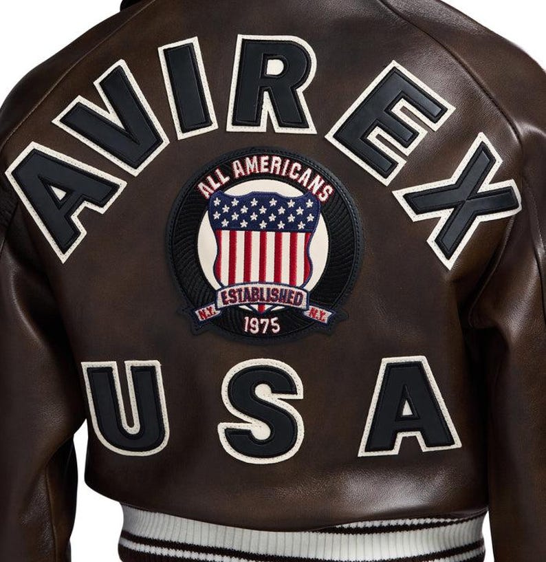 May include: Brown leather jacket with the word "AVIREX" across the shoulders in black and white lettering. A circular patch with the words "ALL AMERICANS" and "ESTABLISHED 1975 N.Y." is centered below the lettering. The word "USA" is below the patch.