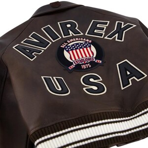 May include: Brown leather jacket with the word "AVIREX" in large white letters outlined in black. A patch with the American flag and the words "ALL AMERICANS ESTABLISHED 1975 N.Y." is centered. The word "USA" is below the patch.