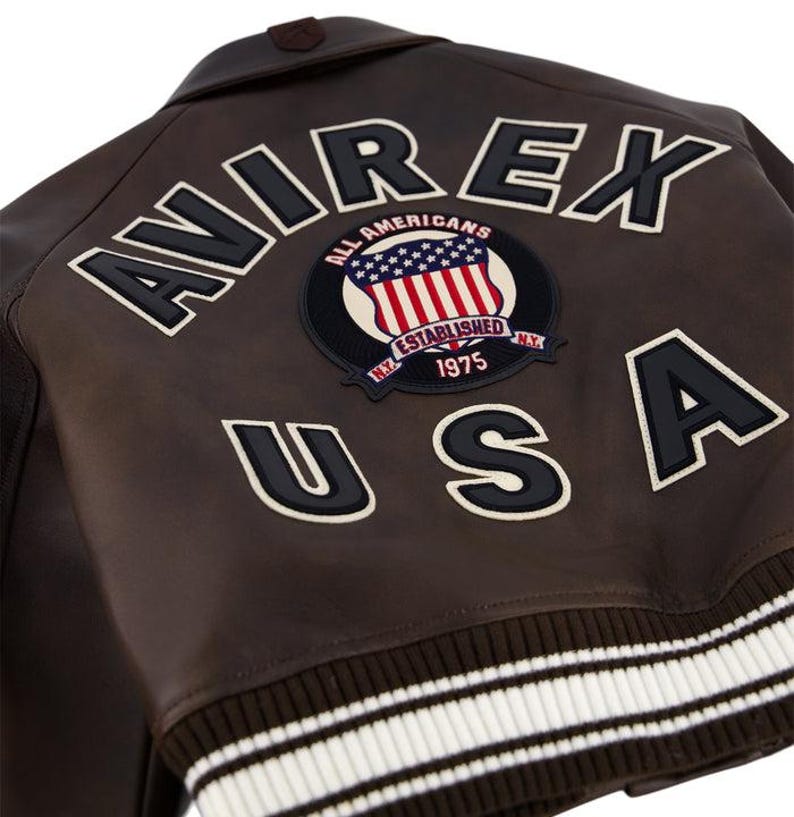 May include: Brown leather jacket with the word "AVIREX" in large white letters outlined in black. A patch with the American flag and the words "ALL AMERICANS ESTABLISHED 1975 N.Y." is centered. The word "USA" is below the patch.