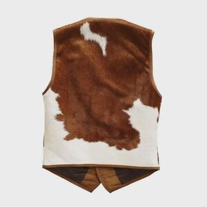 May include: A brown and white cowhide vest with a V-neck and a two-button closure at the waist. The vest features a brown leather trim and a brown suede back panel. The vest is a western-style garment.
