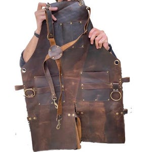 Könnte beinhalten: Eine braune Lederschürze mit mehreren Taschen und Riemen. Die Schürze hat eine Messing-Schnalle und einen Messing-Ring an der Seite.