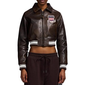 May include: A brown leather cropped jacket with a zip-up front, collar, and ribbed cuffs and hem. The cuffs and hem feature white stripes. A patch with text and a crest is on the upper left chest.