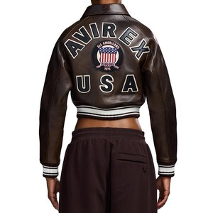 May include: A brown leather jacket with the text "AVIREX" and "USA" on the back. The jacket has a white and black striped trim on the cuffs and waist. A patch with the words "ALL AMERICANS" and "ESTABLISHED 1975" is also visible.