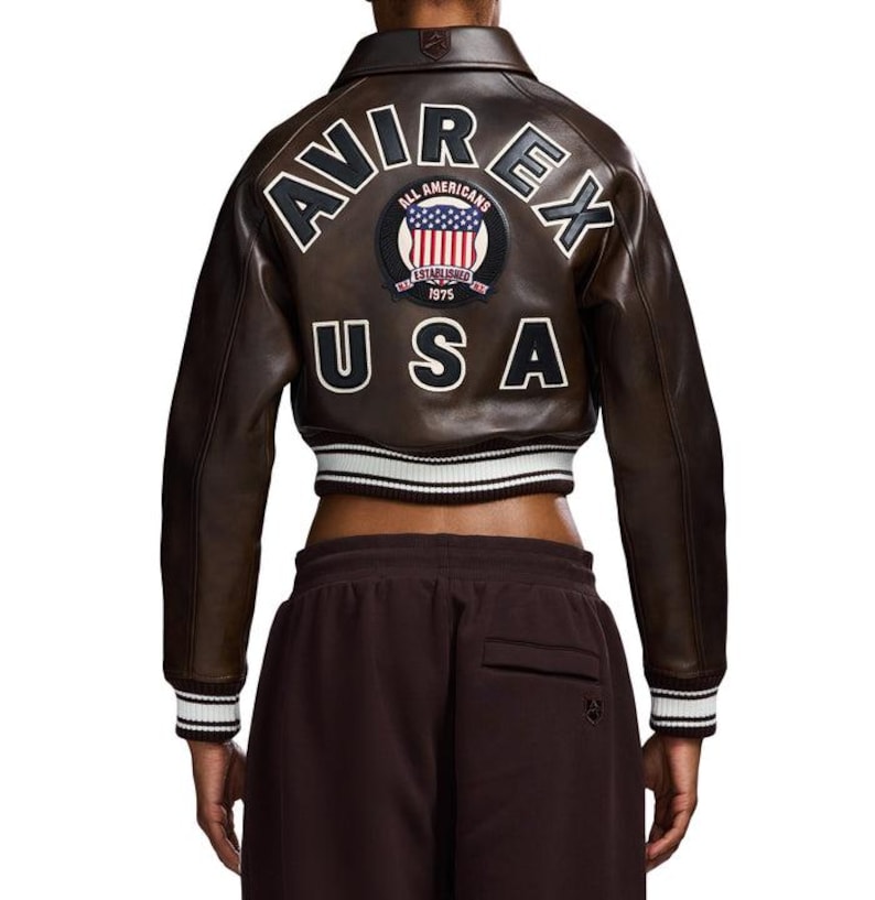 May include: A brown leather jacket with the text "AVIREX" and "USA" on the back. The jacket has a white and black striped trim on the cuffs and waist. A patch with the words "ALL AMERICANS" and "ESTABLISHED 1975" is also visible.