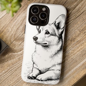 May include: A white phone case featuring a detailed black and white illustration of a Corgi dog. The case is on a wooden surface, with a camera and greenery in the background. The dog is depicted in a seated position, looking to the side.