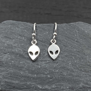May include: A pair of silver alien head dangle earrings. The earrings are small and delicate, and they feature a simple alien head design.
