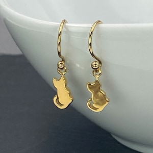 May include: A pair of gold dangle earrings featuring a small cat silhouette charm. The earrings have a simple, minimalist design.