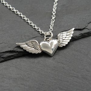 May include: A silver necklace with a heart-shaped pendant and attached wings. The heart is polished, and the wings have detailed feathering. The chain is a simple link style. The pendant hangs against a dark background.