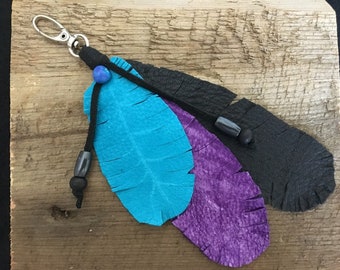 May include: A handmade keychain featuring three leather feathers in turquoise, purple, and black. The feathers are attached to a black cord with beads and a silver-tone clasp. The background is a weathered wooden surface.