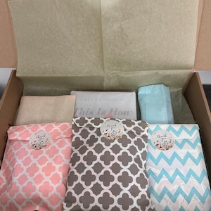 Op de afbeelding: Een open kartonnen doos gevuld met verpakte pakketten. De pakketten zijn in verschillende kleuren en patronen, waaronder roze, grijs en blauw. De pakketten zijn verzegeld met stickers met de tekst "Thank you". Er is ook een boek met de titel "This Is How".