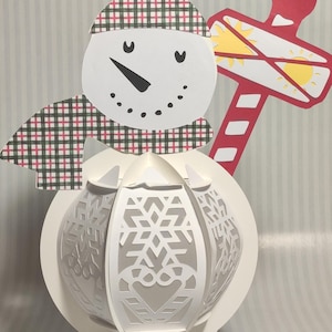 May include: A white papercraft snowman with a red and white striped candy cane, a red and white plaid hat, and a green and white plaid scarf. The snowman's body is a sphere with a snowflake design.