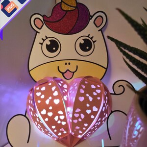 Può includere: Un unicorno bianco con un corno rosa e dorato che tiene una lampada rosa a forma di cuore con pois bianchi. La lampada è illuminata da luci bianche.