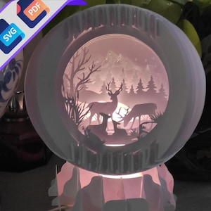 Może przedstawiać: Biała lampa z papieru z jeleniem i leśną sceną w środku. Lampa jest oświetlona od dołu, rzucając ciepłe światło na scenę.