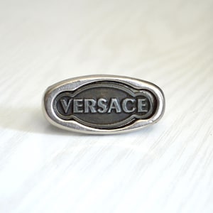 May include: A silver-coloured metal ring featuring the embossed word "VERSACE". The ring is rectangular with rounded edges and a dark grey interior.
