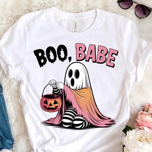 Peut inclure: T-shirt blanc avec un fantôme de dessin animé tenant un seau de citrouille. Le fantôme porte une cape rose et orange, avec des jambes rayées noires et blanches. Le texte "BOO, BABE" est imprimé au-dessus du fantôme en noir et rose.