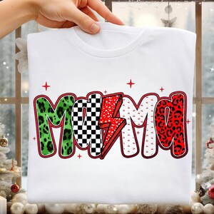 Peut inclure: T-shirt blanc avec le mot "MAMA" dans un design coloré. Les lettres présentent un imprimé léopard vert, un motif à carreaux noir et blanc, un éclair rouge et un imprimé léopard rouge. Des croix rouges sont dispersées autour du mot.