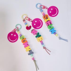 May include: Three colorful beaded keychains with pink smiley face charms and personalized names: Sunny, Jihye, and Sieun. Each keychain features a mix of bright colors and a knotted tassel.
