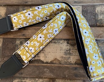 Puede incluir: Una correa de guitarra amarilla con un patrón floral blanco. La correa tiene extremos de cuero negro y un detalle de costura blanca.