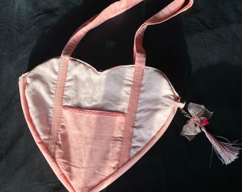Peut inclure: Un sac à main en forme de cœur rose avec une fermeture éclair et une petite poche sur le devant. Le sac a une longue bandoulière et un charme en pompon rose.