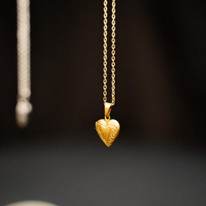 Puede incluir: Un colgante de corazón de oro cuelga de una delicada cadena de oro. El medallón tiene un diseño detallado y es una joya clásica.