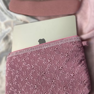 Könnte beinhalten: Eine rosa Laptop-Hülle mit einem zarten floralen Spitzenmuster und weißem Besatz. Die Hülle ist so konzipiert, dass sie einen Laptop vor Kratzern und Stößen schützt.