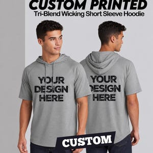 May include: A heather grey short-sleeve hoodie featuring a hood with drawstrings and raglan sleeves. The front and back display the black text "YOUR DESIGN HERE" in a distressed font. The top text reads "CUSTOM PRINTED Tri-Blend Wicking Short Sleeve Hoodie".