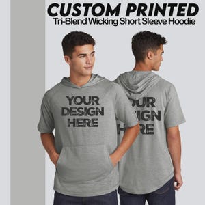 Op de afbeelding: Grijze tri-blend wicking hoodie met korte mouwen, een voorzak en een capuchon. Op de voorkant van het shirt staat "YOUR DESIGN HERE" in zwarte blokletters. De achterkant van het shirt leest "YOUR DESIGN HERE".