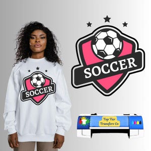 May include: White sweatshirt with a soccer-themed graphic. The design features a soccer ball inside a pink shield with the word "SOCCER" in a black banner. Three black stars are above the shield. A printer with the text "Top Tier Transfers Co" is in the image.