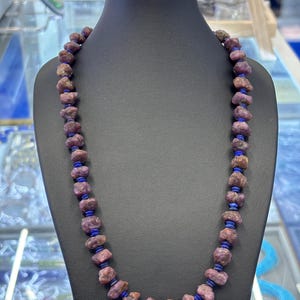 May include: A necklace of rough-cut, reddish-purple gemstones, with small, dark blue beads. The necklace is displayed on a black mannequin, highlighting the jewelry's design and colour contrast. The gemstones have a natural, irregular shape.