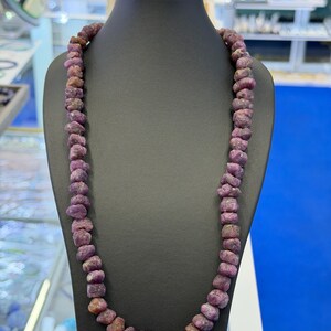 May include: A necklace of raw, uncut ruby gemstones. The stones are a deep, rich purple colour, strung together to form a long strand. The necklace is displayed on a black mannequin.