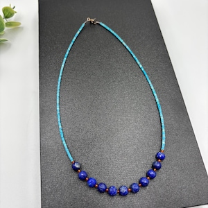May include: A delicate necklace featuring small, turquoise-colored beads strung together with larger, faceted, deep blue beads. The necklace has a gold-colored clasp and is displayed on a black surface. The necklace is a piece of jewelry.