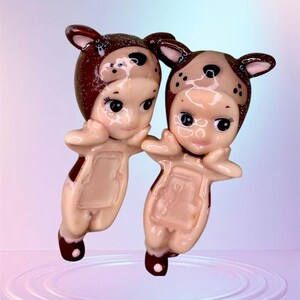 Pu&ograve; includere: Due figurine in ceramica di bambini vestiti con costumi da cucciolo marroni e bianchi. Le figurine sono in piedi su uno sfondo blu chiaro con un effetto increspato.