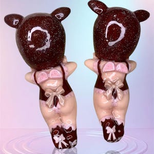 Puede incluir: Dos figuras de cerdos marrones y rosas con detalles de purpurina. Los cerdos llevan lencería rosa y blanca con lazos. Las figuras están de pie sobre una superficie reflectante.