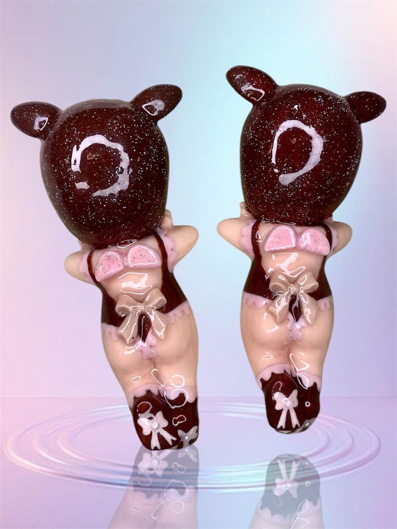 Pu&ograve; includere: Due figurine di maiale marroni e rosa con accenti di glitter. I maiali indossano lingerie rosa e bianca con fiocchi. Le figurine sono in piedi su una superficie riflettente.